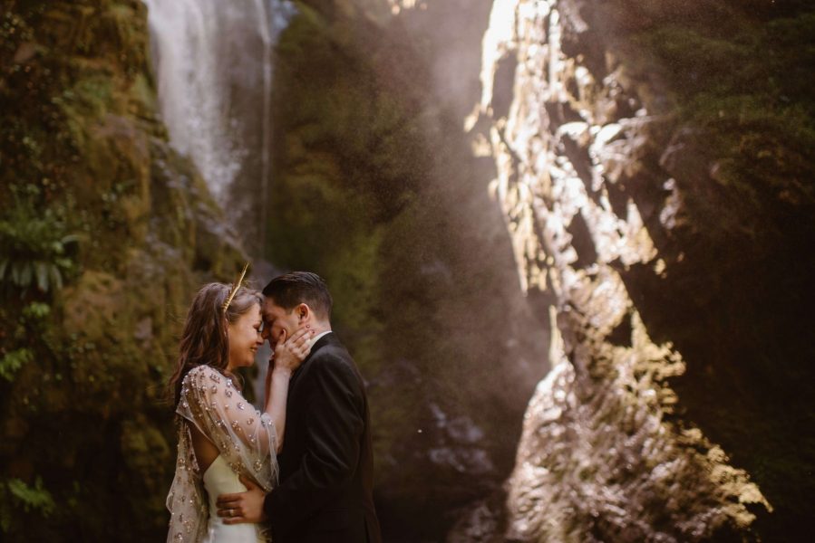 A bride, wearing a crown, holds her husbands face near an Oregon waterfall.