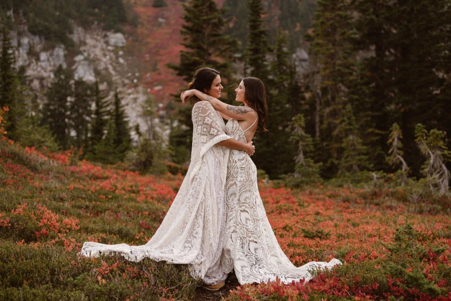 Two brides, dressed in lacey gowns, hold each other. They are flanked by bright orange and red fall colors.