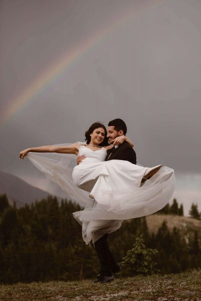 A groom holds his bride in his arms and twirls around. A bright Colorado rainbow sits behind them.