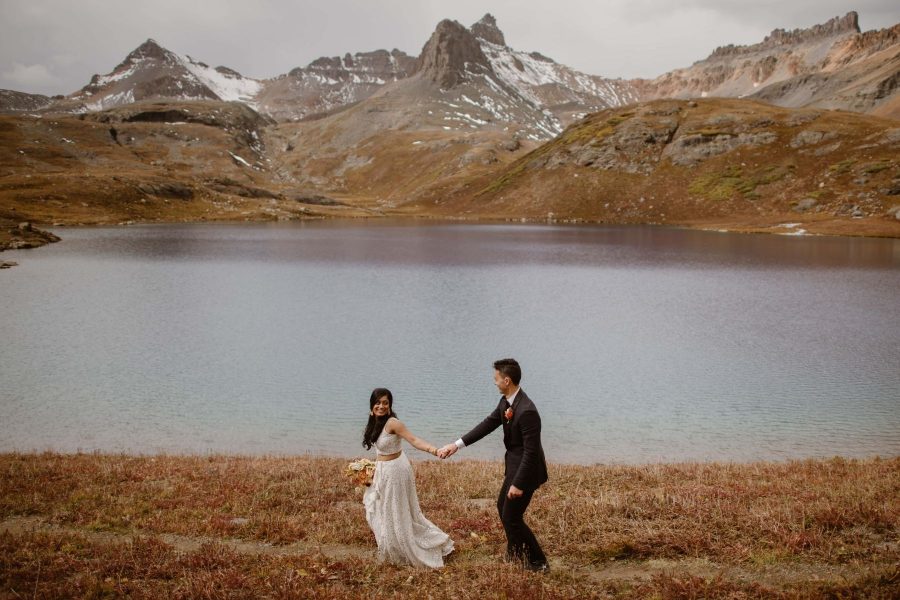 A bride and groom run on the grass in front of an icy lake in Colorado