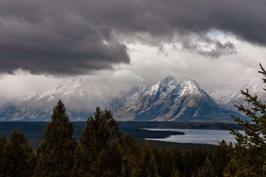 The sun peeks through stormy clouds above the Tetons, illuminating a couple mountains in the distance.