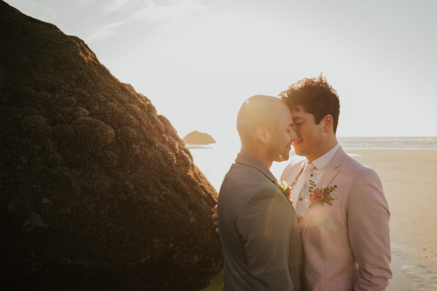 Two Grooms embrace on a beach. There is a large rock beside the. One is wearing a gray suit. One is wearing a pink suit.