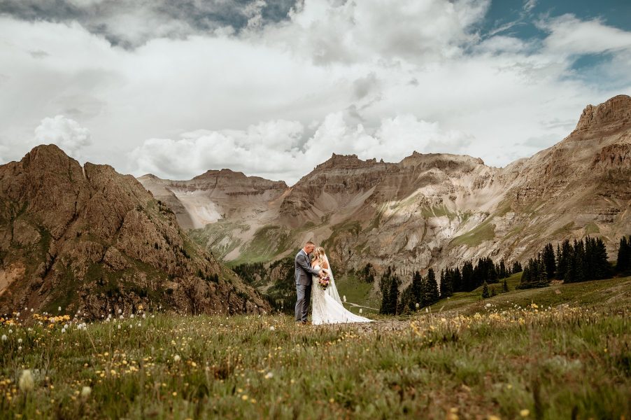 Elopement couple stand surrounded by blue skies, far off peaks, and wildflowers as far as the eyes can see during their elopement in the San Juan mountains