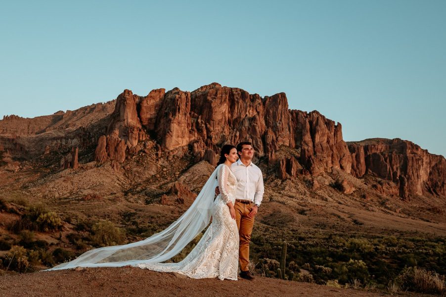 bride and groom look off into the sunset as her long cape billows behind her in the Superstition Mountains