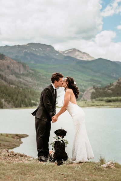 couple holds hands over the silhouette of their dog and kiss with snow capped Colorado peaks framing them in the distance