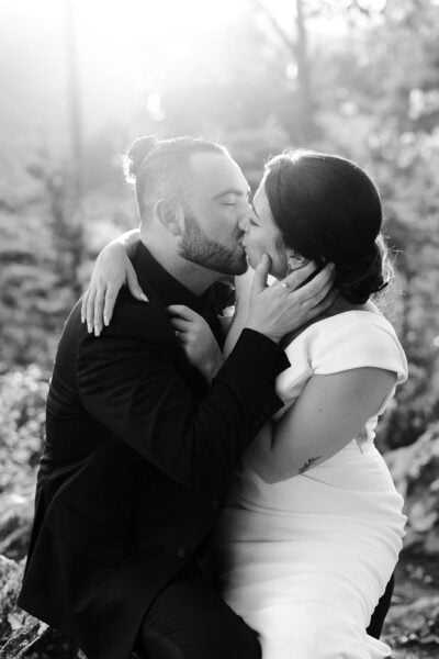 A couple in formal attire share a passionate kiss outdoors. The man, in a suit, has a neatly trimmed beard and hair pulled back in a bun. The woman, in a white dress, gently holds his face. Sunlight filters through the trees in the background. Black and white image by Erin Morrison Photography.