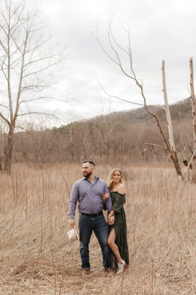 A man and woman stand in a dry grassy field with bare trees. The man wears a gray shirt and jeans, holding a white jug. The woman, in a green off-the-shoulder dress, holds his arm. Overcast sky and distant hills form the background. Photo by Smoky Mountain Engagement Photographer Erin Morrison Photography.