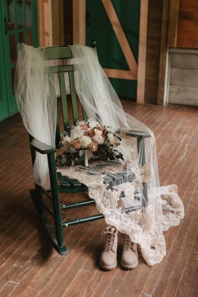 A green rocking chair holds a delicate lace veil and bouquet of white and blush flowers. A pair of tan boots rests on the wooden floor beside the chair, creating a rustic and romantic scene in a warmly lit room. Photo by Smokies Elopement Photographer Erin Morrison.