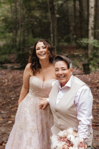 A happy couple stands together on a forested path. One person wears a white dress with lace details, while the other wears a light beige vest and white shirt. Both are smiling, and the scene is filled with fallen leaves and trees in the background. Photo by LGBTQIA+ Friendly Photographer Erin Morrison Photography.