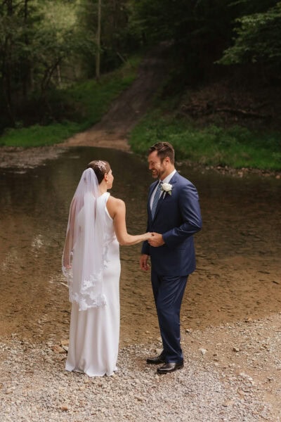 A bride in a white dress and veil stands holding hands with a man in a blue suit by a shallow river in a forested area, conveying a serene and intimate moment. Photographer by Erin Morrison Photography.