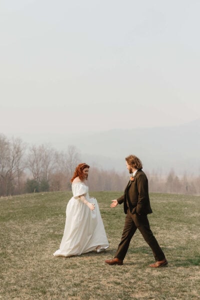 A red-haired woman in a white dress and a man in a dark suit walk towards each other on a grassy field. The background features a misty landscape with barren trees and hazy Smoky Mountains. Photo by Elopement Photographer Erin Morrison.