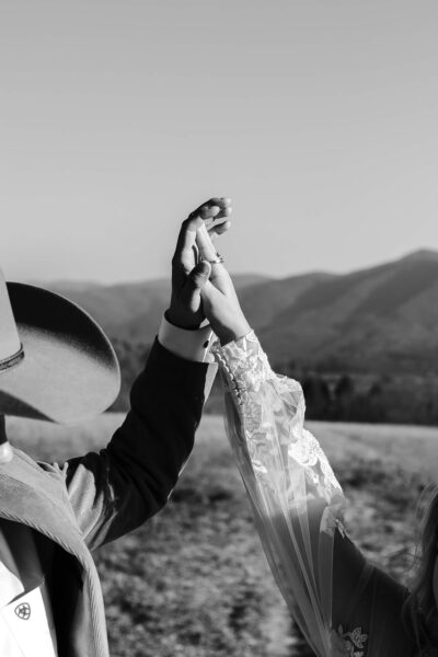 A couple holding hands in a field, wearing a cowboy hat and a lace sleeve. The black-and-white photo captures the Smoky Mountain landscape in the background. Photo by Cades Cove Photographer Erin Morrison Photography.
