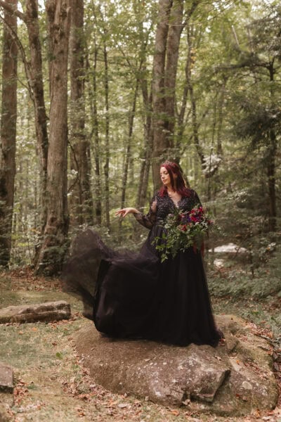 A woman in a long, flowing black gown stands on a rock in a forest, surrounded by tall trees. She holds a bouquet of dark flowers and her red hair contrasts with the lush green background. Her dress drapes elegantly over the rock at the Appalachian Clubhouse. Photography by Erin Morrison Photography.