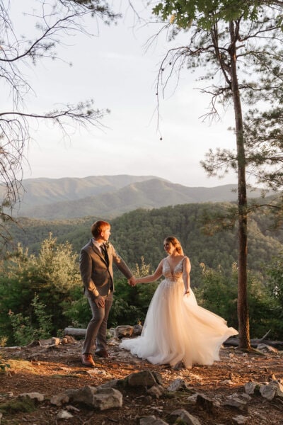 A couple in wedding attire holds hands on a rocky path, surrounded by trees, with a scenic mountain range in the background under a clear sky. The bride wears a flowing gown, and the groom is in a suit, both bathed in warm sunlight. Photo by Smoky Mountain Photographer Erin Morrison.