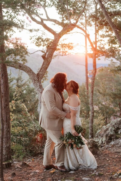 A couple in wedding attire shares a kiss in a forest at sunset. The bride holds a bouquet and wears a lace dress, while the groom wears a beige suit. Sunlight filters through the trees, creating a warm, romantic atmosphere at The Magnolia Venue. Photographed by Erin Morrison.