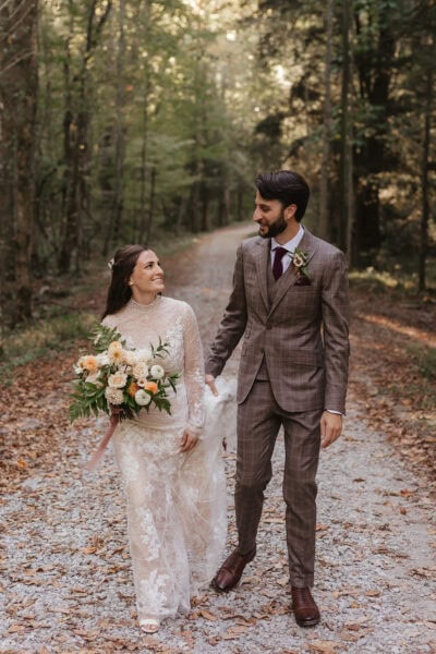 A bride and groom walk hand in hand on a forest path in Elkmont. The bride wears a lace wedding dress and carries a bouquet of flowers. The groom is in a brown suit with a vest. They are smiling at each other, surrounded by tall trees and fallen leaves. Photo by Erin Morrison Photography.