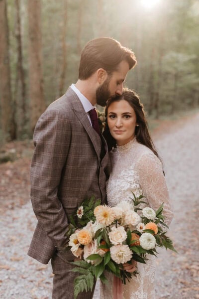 A couple stands on a forest path. The man, in a plaid suit, gently kisses the woman’s forehead. She wears a lace dress and holds a bouquet of pale flowers. Sunlight filters through the trees in the background at Elkmont Elopement. Photo by Erin Morrison Photography.