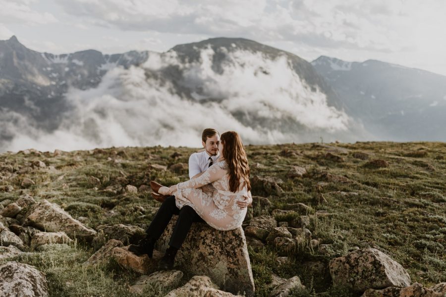 couple in wedding attire sitting on rock and cuddling with mountains and clouds in the background