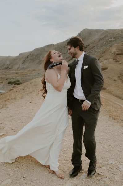 couple in wedding attire laughing on top of windy mountain