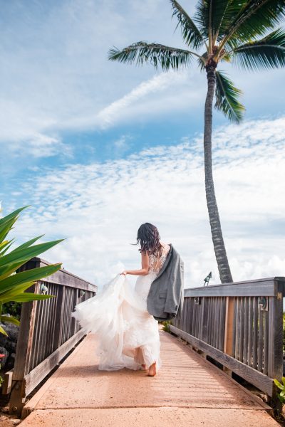 Bride strolling under a palm tree in her wedding dress with her groom's jacket thrown over her shoulder in Kauai, Hawaii