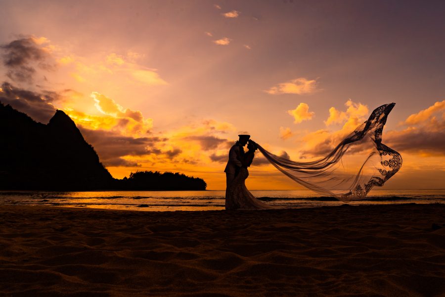 Bride and groom silhouetted in the sunset with the bride's veil blowing in the breeze in Kauai, Hawaii