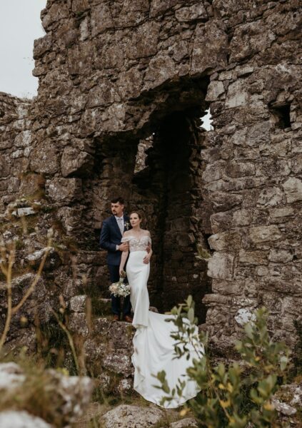 A bride and groom stand close together at an abandoned castle in Ireland.