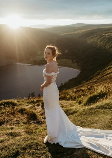 A woman in a wedding dress looks at the camera. She is standing next to a mountain lake in Ireland.
