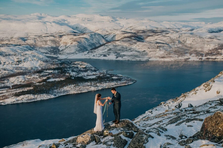 Bride and groom performing handfasting ceremony on a mountaintop among snow-capped mountains in Alta Norway after reading their elopement vows to each other