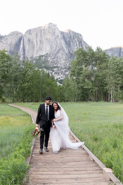 Yosemite Elopement Bridal couple walking the boardwalk trail with the waterfall behind them.