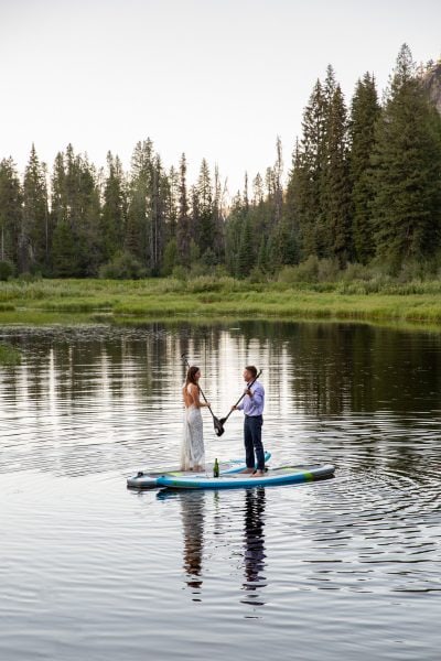 Bride and groom stand on paddleboards in the middle of a lake with their paddles touching between them.