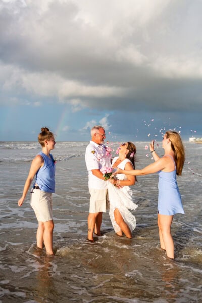 A bride and groom stand in the Gulf of Mexico laughing as their daughters throw flower petals at them.
