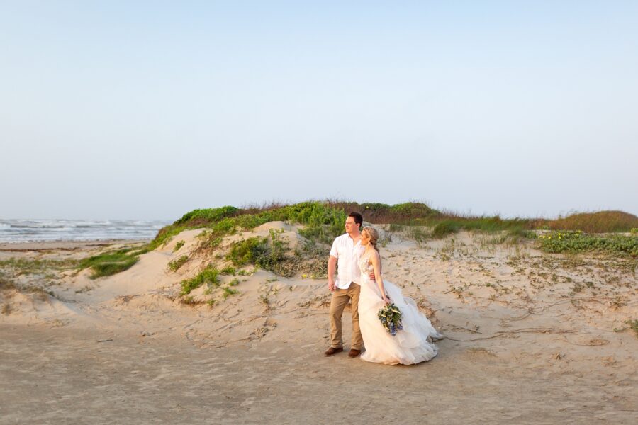 A bride and groom stand in front of a large sand dune looking out at the sunrise over the ocean in Galveston Texas.