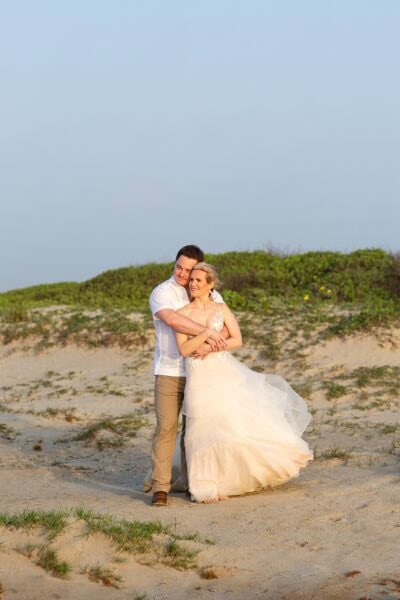 A groom embraces his bride in front of a large sand dune looking out at the sunrise over the ocean in Galveston Texas.