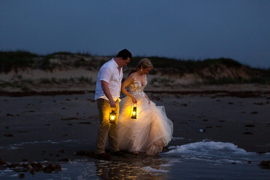 A bride and groom holding lanterns play at the water's edge during the sunrise blue hour.
