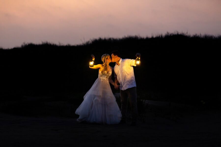 A bride and groom holding lanterns kiss in front of a large sand dune during the sunrise blue hour.