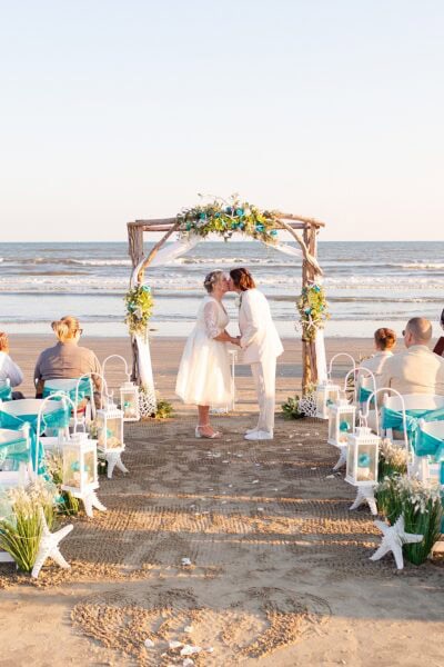 Two brides kiss under a wedding arch on Galveston Beach at their wedding ceremony.