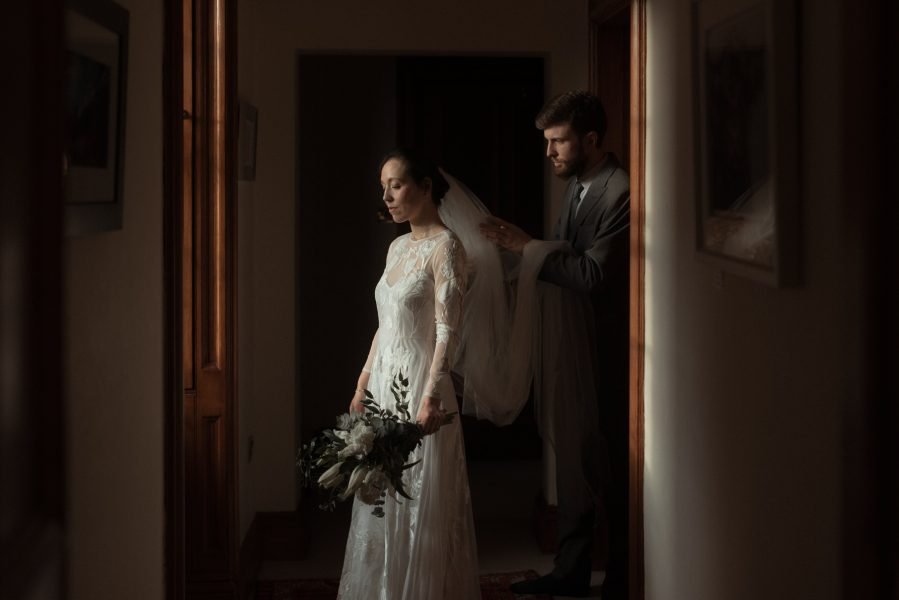 Bride and groom standing in a traditional wood panelled room, while the groom helps her to attach her veil.