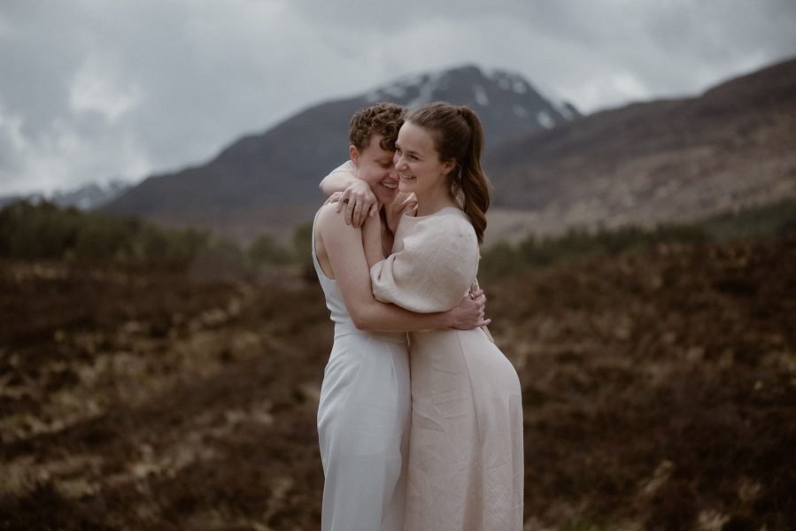 Two brides embracing in front of a mountain backdrop in Scotland