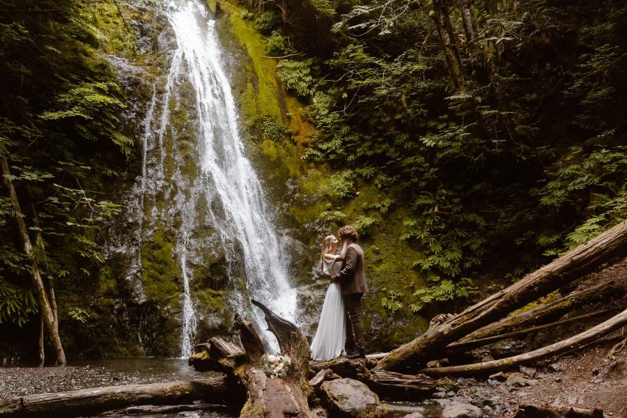 washington-waterfall-elopement-couple-in-wedding-attire-stand-on-a-log-in-front-of-a-waterfall
