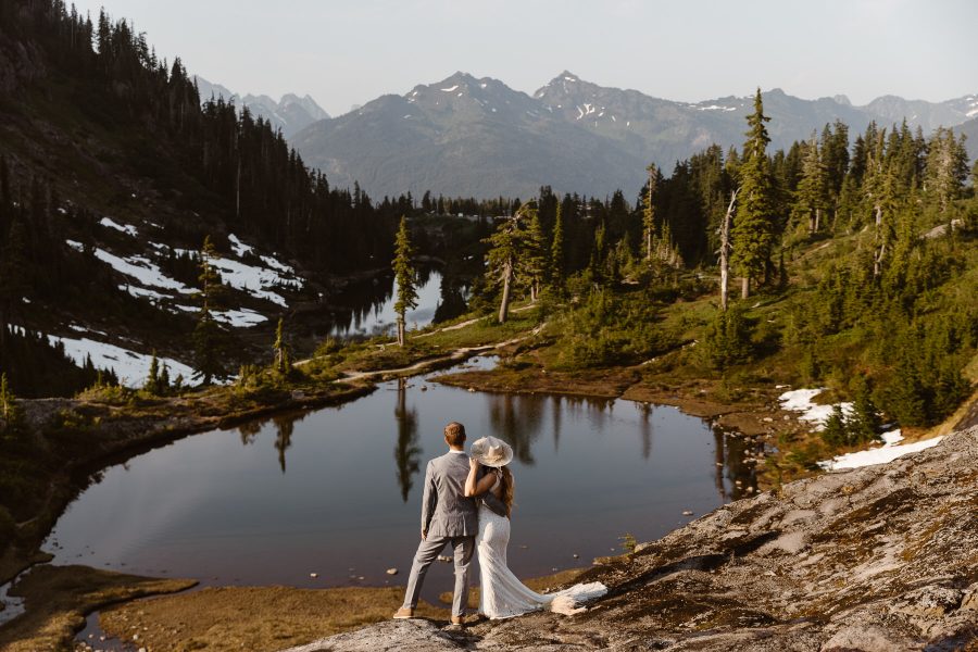 couple-in-wedding-attire-stands-in-front-of-a-lake-overlooking-mountains-at-their-north-cascades-elopement