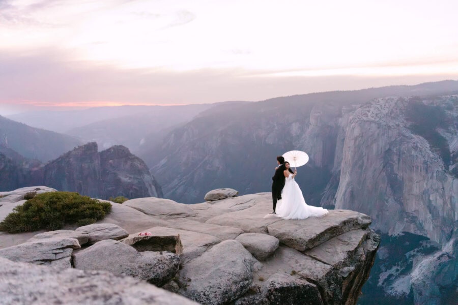 Bride and groom standing on the cliffs of Taft Point in Yosemite at sunset, showcasing breathtaking views of the valley. Bride holding a parasol.