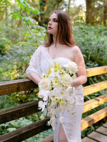 Bride standing gracefully on a rustic bridge at Ecola State Park, holding her bouquet, surrounded by lush greenery and serene forest views.
