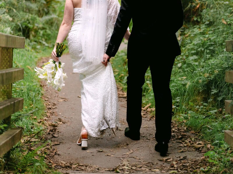 Groom helping the bride hold her dress as they hike a forest trail in Ecola State Park, with their hiking shoes visible on the path.
