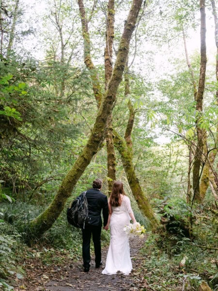 Couple walking hand in hand on a trail in Ecola State Park, captured from behind with towering trees and a lush forest stretching ahead.