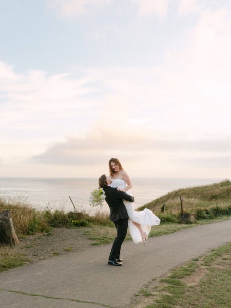 Groom lifting and twirling the bride at the overlook point in Ecola State Park, with sweeping coastal views and dramatic cliffs in the background.