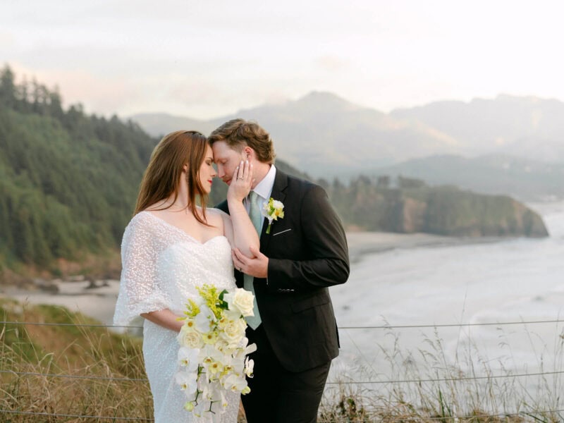 Bride and groom sharing a romantic embrace at the overlook with Haystack Rock in the background, the bride gently holding the groom's face in one hand and her bouquet in the other.
