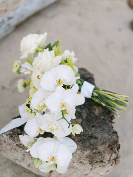 A close-up of a white orchid and rose bouquet resting on a rustic tree stump against the soft sand at Indian Beach.