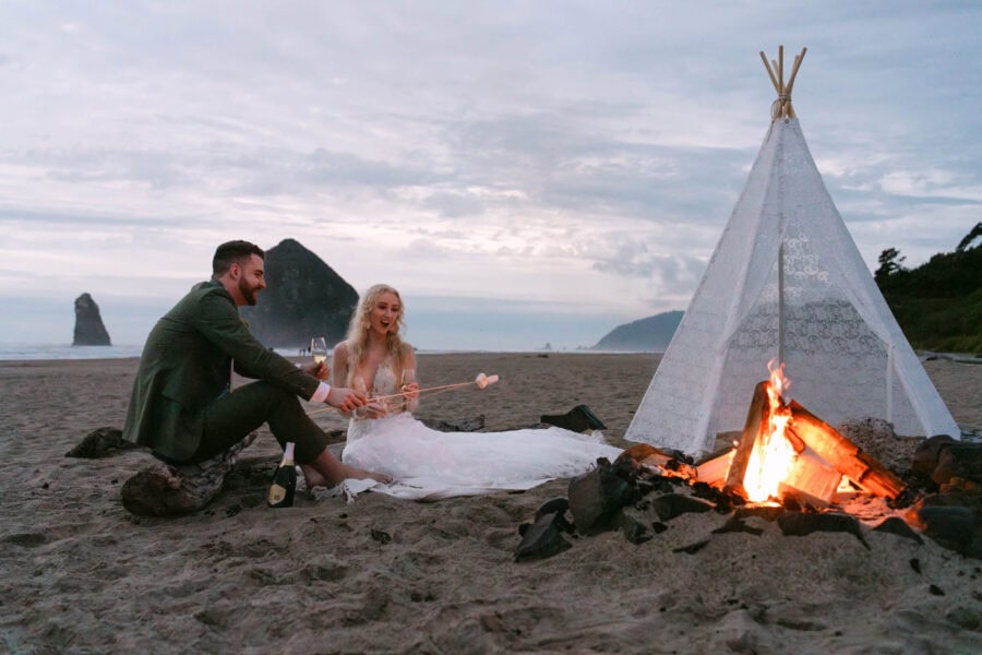 Bride and groom roasting marshmallows over a campfire at Cannon Beach, with a white teepee and Haystack Rock in the background.