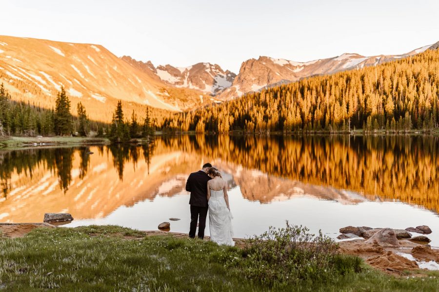Newlyweds face a still lake, reflecting a fiery sunset across pine trees and snow-capped mountains