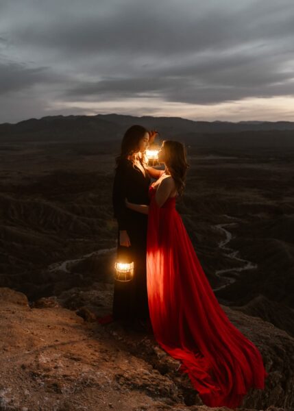 Two women in elegant dresses, one in black and one in red, stand on a rocky overlook at twilight, holding a lantern that illuminates their faces against a dramatic backdrop of dark, cloud-covered skies and the rugged terrain of a canyon below